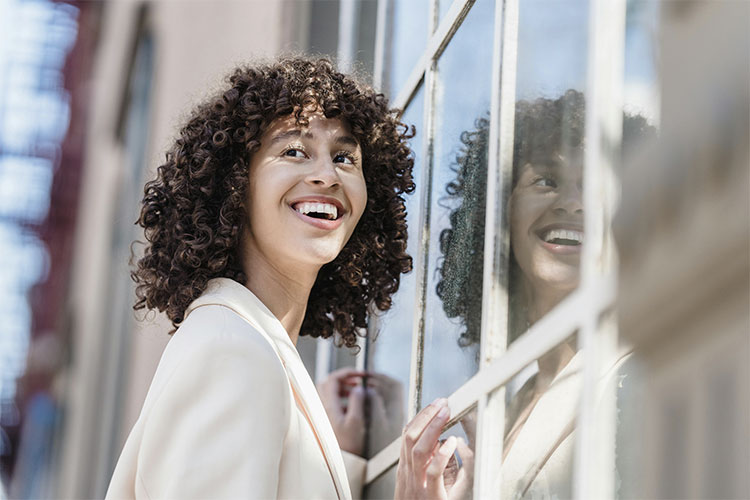 Smiling woman standing next to a closed glass door, represents the barriers we face on the road to reinvention and how therapy can help us overcome them.