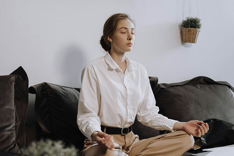 Woman sitting on a couch in her office focusing on her breathing, represents how you can put the theory of nervous system regulation into practice.